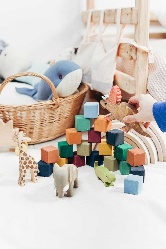 creative A childs hand arranges colorful wooden toys and animal figures on a playroom floor.